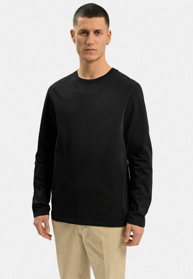 Svart långärmad t-shirt i bomull, med rundad halsringning och en subtil logotyp på bröstet, i kombination med ljusa byxor.