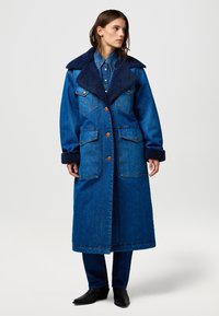 Płaszcz dżinsowy w średnim odcieniu niebieskiego z dużym granatowym kołnierzem z futerka syntetycznego, zapięciem na guziki, podwójnymi kieszeniami na piersi i długimi rękawami. Posiada kontrastowe przeszycia.