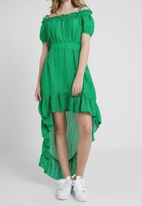 Vihreä olkaimeton mekko, jossa on teksturoitu kuvio, röyhelöreuna ja linnankurkkuna; yhdistettynä valkoisiin tennareihin rentoon ilmeeseen.