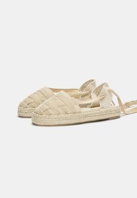 Bežové espadrilky s pleteným plátnem, kulatou špičkou a křížovými popruhy. Podešev obalená jutou dodává texturu. Lehce provedení pro pohodlí.