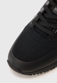 Schwarze Sneakers mit strukturiertem, atmungsaktivem Mesh-Obermaterial, glattem Schutz-Kappen und schwarzen Schnürsenkeln vor einem weißen Hintergrund.