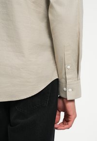 Camicia beige a maniche lunghe con finitura testurizzata, caratterizzata da due bottoni bianchi sul polsino e un orlo curvo; abbinata a pantaloni di denim neri.