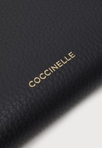 Black textured leather wallet featuring the gold embossed brand name "Coccinelle" on the front. Smooth surface with prominent grain detail.