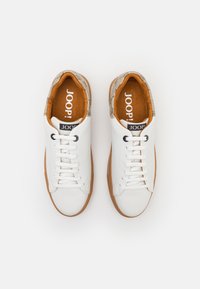 Zapatillas blancas con una parte superior de cuero liso, suela de goma marrón y un acento de tela marrón texturizada en el cuello. Logo negro visible.