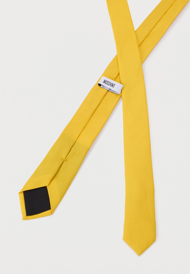 Cravate en soie jaune avec une texture lisse. Elle présente une extrémité pointue et une doublure intérieure noire. Étiquette visible avec le nom de la marque "Moschino".