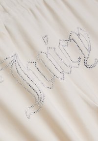 Tissu crème avec des embellissements en strass argentés brillants formant des motifs fluides et ondulés sur toute la surface. Texture douce, taille élastique.