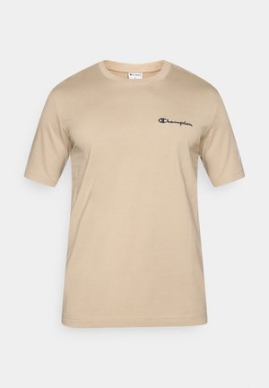 Camiseta de algodón beige con mangas cortas. Presenta un logo "Champion" bordado en azul marino en el pecho izquierdo y un cuello redondo acanalado.