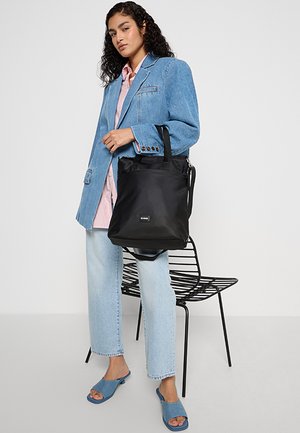 Donna che indossa un blazer di denim blu e jeans, una camicia rosa, sandali blu, tiene una borsa a tracolla nera, in piedi accanto a una sedia di metallo nera.