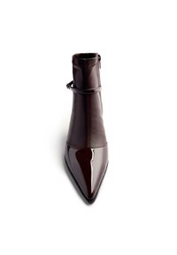 Bottine en cuir verni marron avec un bout pointu, fermeture éclair sur le côté et une sangle autour de la cheville. Texture lisse avec une finition brillante.