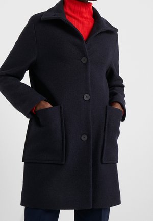 Zwarte wollen jas met drie knopen en grote zakken aan de voorkant, gedragen over een rood geribd coltrui.