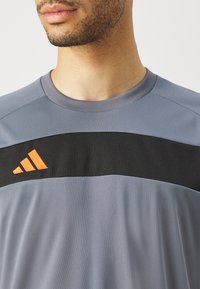 Szara koszulka sportowa z czarnym poziomym paskiem i pomarańczowym logo. Gładka tekstura, krótkie rękawy i okrągły dekolt.