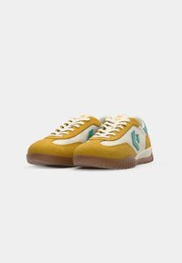 Suede sneakers with a mustard yellow upper, white canvas accents, teal logo detail, and brown rubber sole. Classic lace-up design.