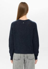 Temno modra volnena jopica s okroglo izrezom in dolgimi rokavi. Ima majhen srebrn gumb na hrbtni strani. V kombinaciji s sivo denim hlačami.