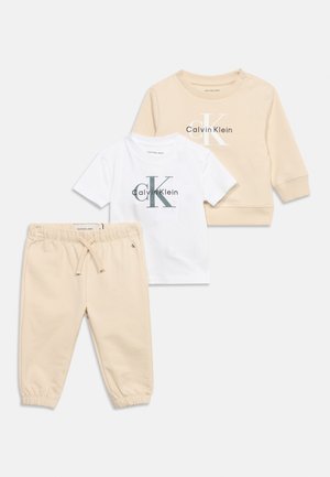 Bež trenirke za malčke, bela kratka majica s kratkimi rokavi Calvin Klein in bež dolga majica s kapuco z logotipom CK, položene ravno na belem ozadju.