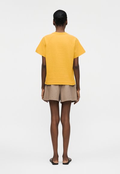 Personne debout de dos, portant une chemise jaune à manches courtes, un short beige et des sandales noires sur un fond clair uni.