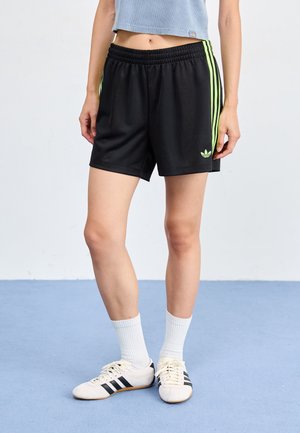 Personne portant un short Adidas noir avec des rayures vertes, des chaussettes blanches et des baskets blanches avec des rayures noires sur un sol bleu.
