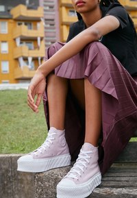 Ljust rosa högstövlar med vita snören och texturerade sulor, parat med en plisserad, djupröd kjol, mot en grönskande bakgrund.