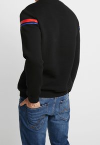 Černá mikina s kulatým výstřihem, s červenými a modrými pruhy na rukávech, doplněná modrými džínami.