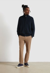 Granatowa, prążkowana dzianinowa kurtka z suwakem, khaki chinosy, granatowe sneakersy z białymi podeszwami. Model stoi z rękami w kieszeniach na jednolitym tle.