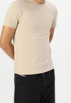 Personne portant un T-shirt beige à manches courtes avec un petit logo sur la poitrine et un pantalon noir, haut du corps visible sur fond uni.