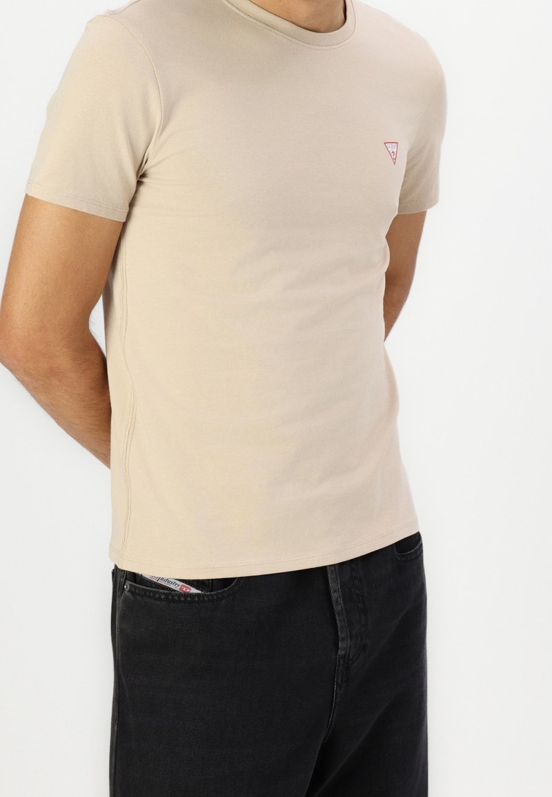 Personne portant un T-shirt beige à manches courtes avec un petit logo sur la poitrine et un pantalon noir, haut du corps visible sur fond uni.