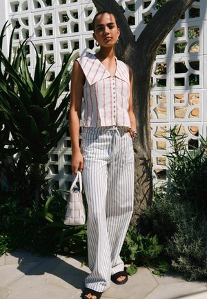 Woman standing outdoors by a tree and white patterned wall, wearing striped sleeveless top and wide-leg striped pants, holding a white handbag.