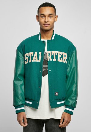 Grüner Varsity-Jacket mit beigem "STARTER"-Schriftzug, Lederärmeln, geripptem Kragen und Bündchen. Getragen über einem hellen T-Shirt und Jeans.