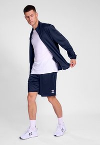 Marineblaue Trainingsjacke mit langen Ärmeln, weißes T-Shirt und marineblaue Shorts; weiße Sportschuhe; Markenlogo auf den Shorts sichtbar.