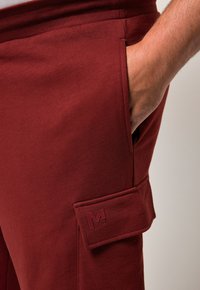 Burgunderfarbene Jogginghosen aus weichem Stoff, mit einer seitlichen Cargo-Tasche mit eingeprägtem "M"-Logo, elastischem Bund und glatter Textur.