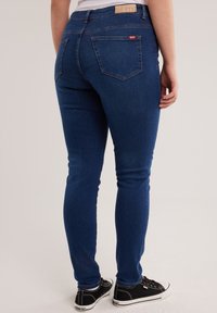 Persoană purtând blugi skinny albastru închis și adidași negri cu talpă joasă, stând cu o mână relaxată pe lângă corp.