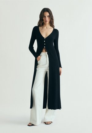 Mujer vestida con un largo cárdigan negro abotonado sobre pantalones blancos de pierna ancha y sandalias negras, de pie contra un fondo claro y liso.