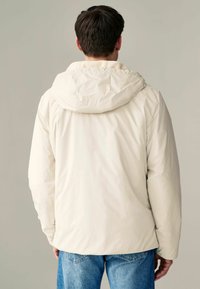 Geacă puffer de culoare bej deschis, cu glugă, având o textură netedă și un șnur ajustabil. Asortată cu blugi din denim albastru.
