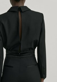 Svart skräddarsydd jumpsuit med långa ärmar, strukturerad krage, dragkedja bak och öppen ryggdetalj. Har en figurnära midja och en stilren design.