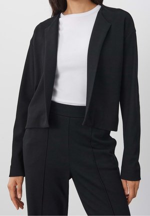 Kvinde iført en sort åben blazer, højtaljede sorte bukser og en hvid ribstrikket top med rund hals mod en ensfarvet baggrund.