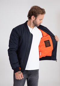 Giacca bomber navy con fodera trapuntata arancione, colletto e polsini a coste, e chiusura con zip. Presenta una patch con il logo all'interno.