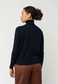 Marineblauwe coltrui met een aansluitend ontwerp, geribbelde onderrand en mouwen, gemaakt van een zachte stof, gezien vanaf de achterkant met een bruine broek.