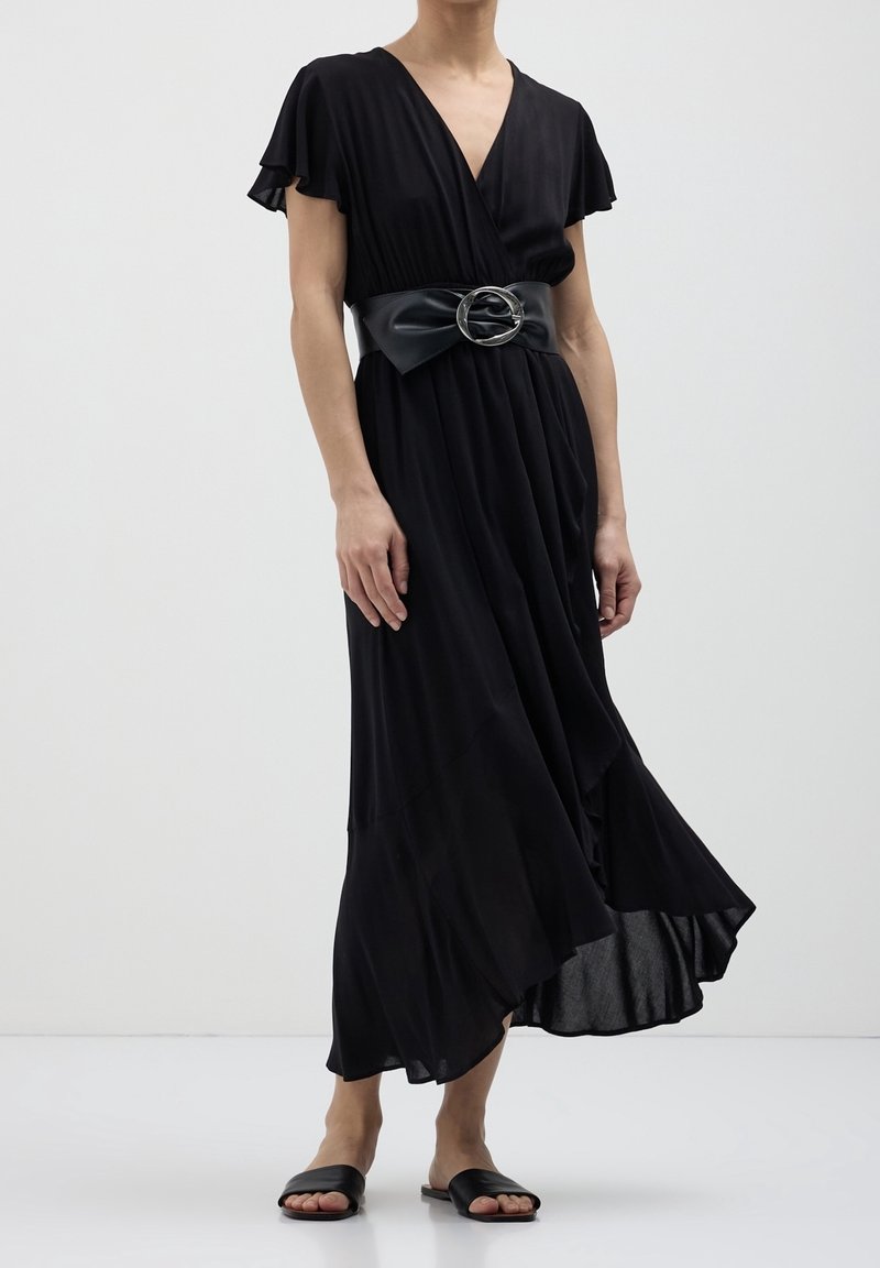 Personne portant une robe portefeuille noire avec des manches papillon et une large ceinture avec une grande boucle ronde, associée à des sandales slide noires.
