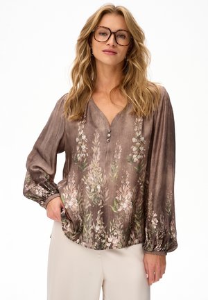 Blondynka w okularach w wzór żółwia i brązowej, kwiecistej, długorękawowej bluzce z guzikami, pozująca z jedną ręką w kieszeni.