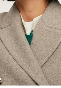 Blazer di lana grigia con un rever affilato, caratterizzato da un unico bottone argentato. Sotto, una camicia verde e bianca con trama appare leggermente.