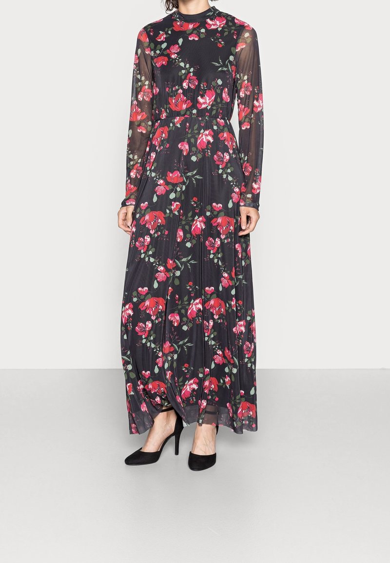 Femme portant une longue robe noire avec des manches longues transparentes à motif floral et des fleurs rouges, associée à des chaussures à talons hauts noires.