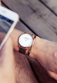 Orologio con cinturino in maglia rosé e quadrante bianco con indicatori neri, accanto a un sottile bracciale dorato con testo inciso.