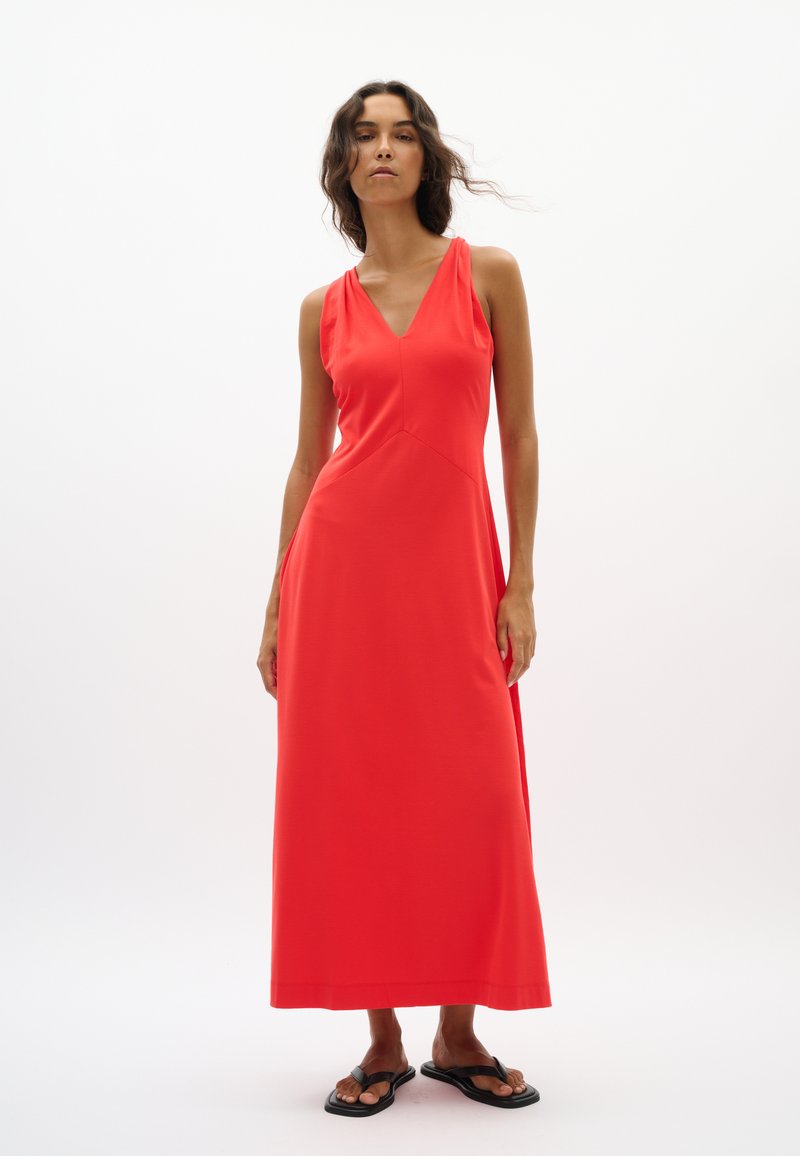 Femme aux longs cheveux foncés portant une robe maxi rouge sans manches avec un col en V et des sandales noires sur un fond blanc uni.