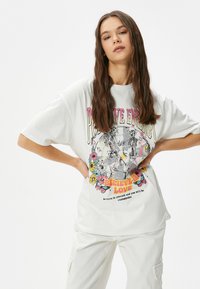 Hvid oversized t-shirt med farverigt grafikdesign, der viser skeletter, blomster og tekst, lavet af blød bomuld med korte ærmer.