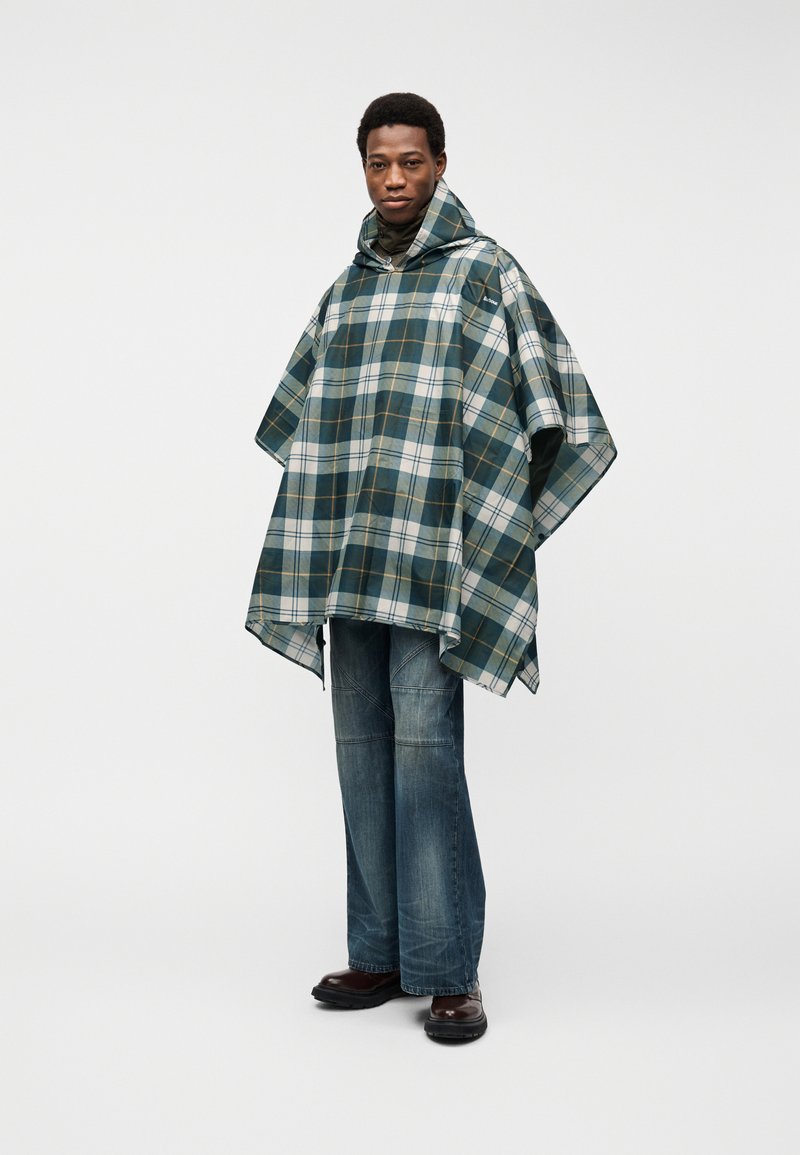 Άνδρας ντυμένος με κουκούλα πόντσο σε πράσινο και άσπρο καρό, μπλε τζιν και σκούρα καφέ παπούτσια, στέκεται μπροστά σε απλό ανοιχτό φόντο.