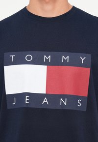 Marineblå bomulds t-shirt med et rektangulært grafisk design med teksten "TOMMY JEANS," en hvid blokk og røde samt blå accenter. Rund hals.