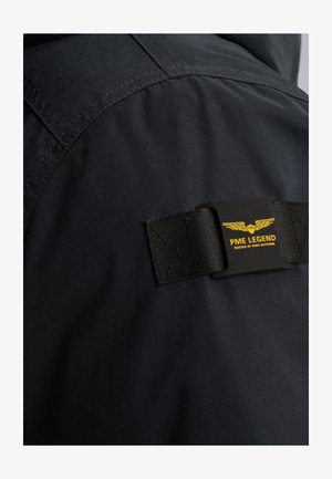 Črna jakna z teksturirano tkanino, z izrazito črno in rumeno oznako "PME LEGEND" in dodano strap detajlom.
