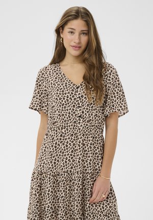 Kobieta o długich brązowych włosach, ubrana w beżowo-czarną sukienkę w panterkę z krótkim rękawem i dekoltem w serek, stojąca na jednolitym tle.