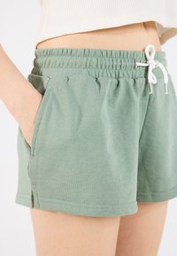 Green cotton shorts with a stretchy waistband, featuring side pockets and a white drawstring. Fabric has a soft texture and casual design.