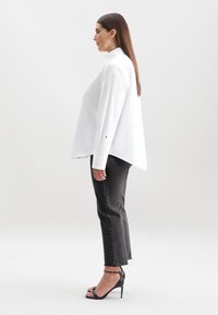 Donna con capelli castani lisci che indossa una camicia bianca oversize, jeans neri a vita alta e sandali neri con tacco alto, in piedi di profilo.