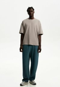 Tricou oversized bej cu mânecă scurtă, asortat cu pantaloni plisati teal și adidași gri cu o suprafață texturată.